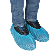 Pair of light blue disposable non-skid SPP shoe covers with printed tread patterns, fitted over black safety work shoes and blue jeans, on a transparent background.