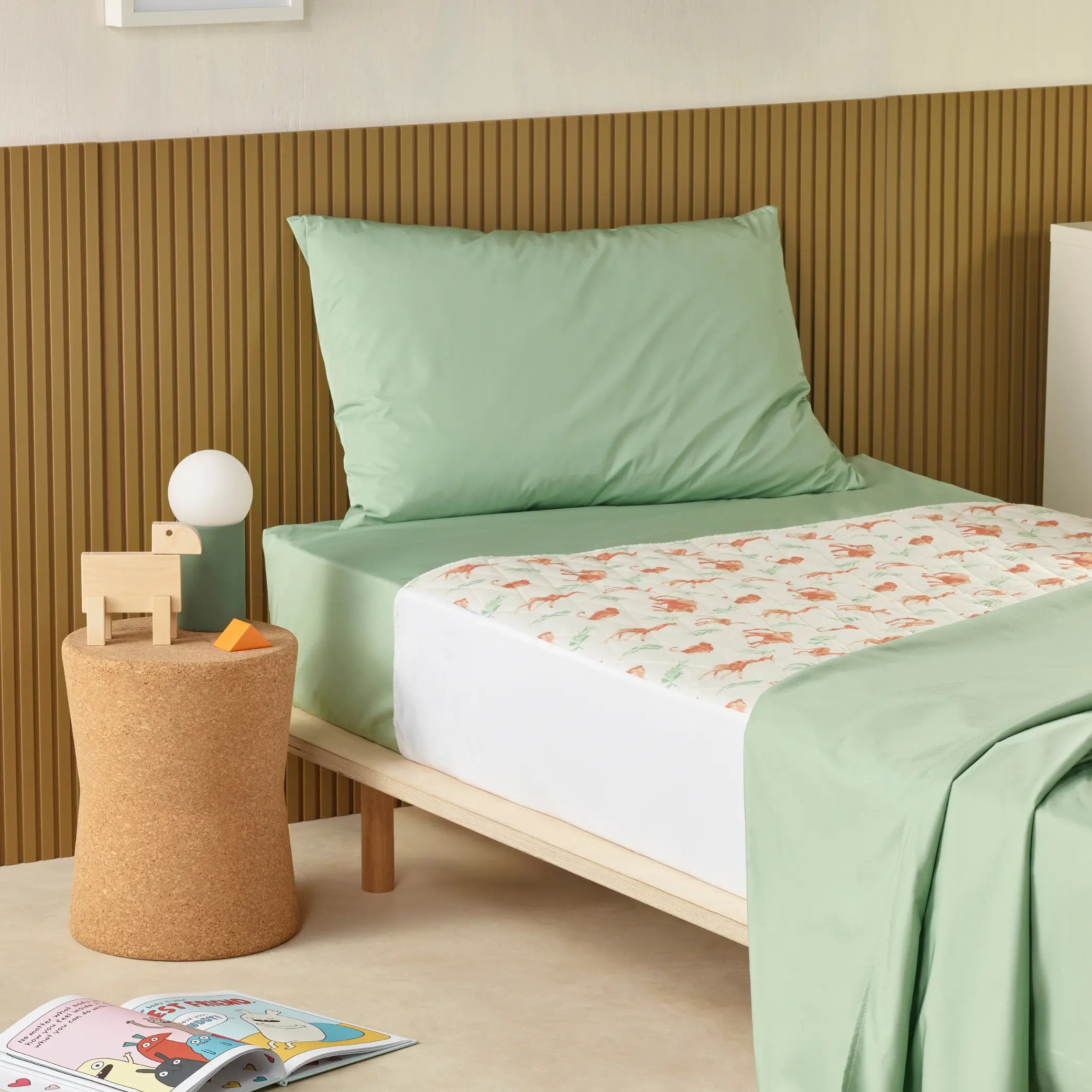 Children’s waterproof absorbent bed pad, also known as kylie bed pad with a playful green animal print, placed on a single bed with sage green bedding. This reusable, leak-proof incontinence pad protects mattresses from accidents, ideal for toilet training and night-time use.