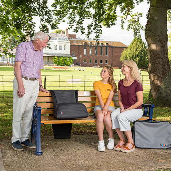 Two women and an older man sitting on a bench with suitcases next to them in a park setting.